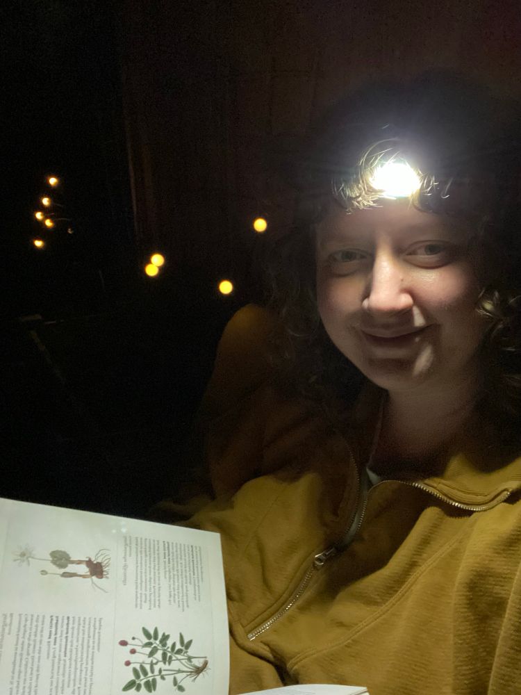 Selfie of me with my headlamp, smiling as I’m holding my her book, in the background is darkness just a few LED fairy lights