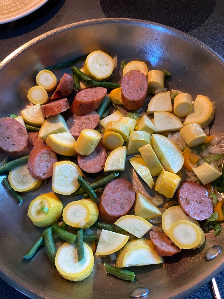 Stainless steel pan with sliced chicken and apple sausage, sliced cook, neck, yellow, summer squash, chopped yard, long or asparagus beans, as well as diced onions and Shishido peppers cooking