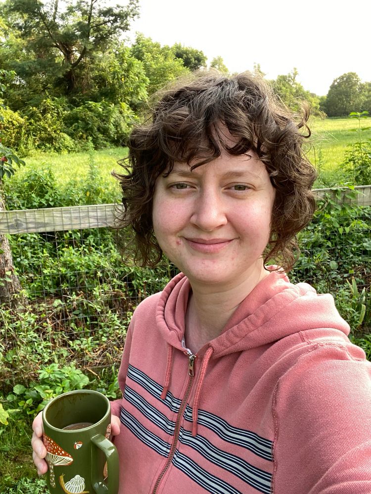 Selfie of me, Soren genderfluid white person with curly brown hair and bangs that’s been pulled back into a ponytail, I’m wearing one of my favorite hoodies faded red with stripes, and holding my favorite mug dark green with woodland creatures on it. Behind me you can See the farmers field and the fence of the property.