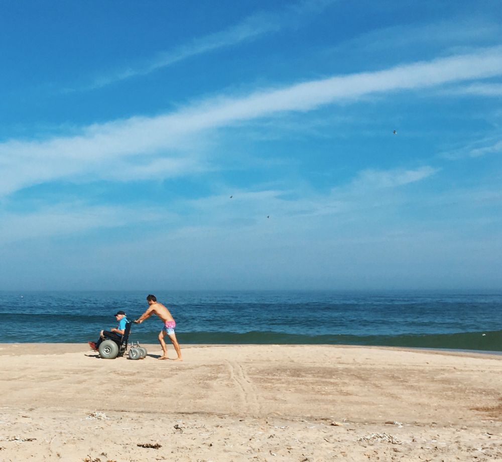A young carer cheerfully pushes an elder in a beach wheelchair, at Nauset Beach, Cape Cod, Massachusetts. 