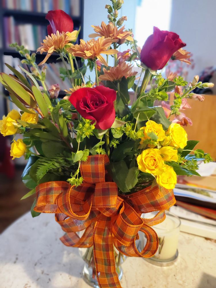 Red and yellow roses with mums and other small flowers in a large glass vase, wrapped in an orange tartan pattern bow
