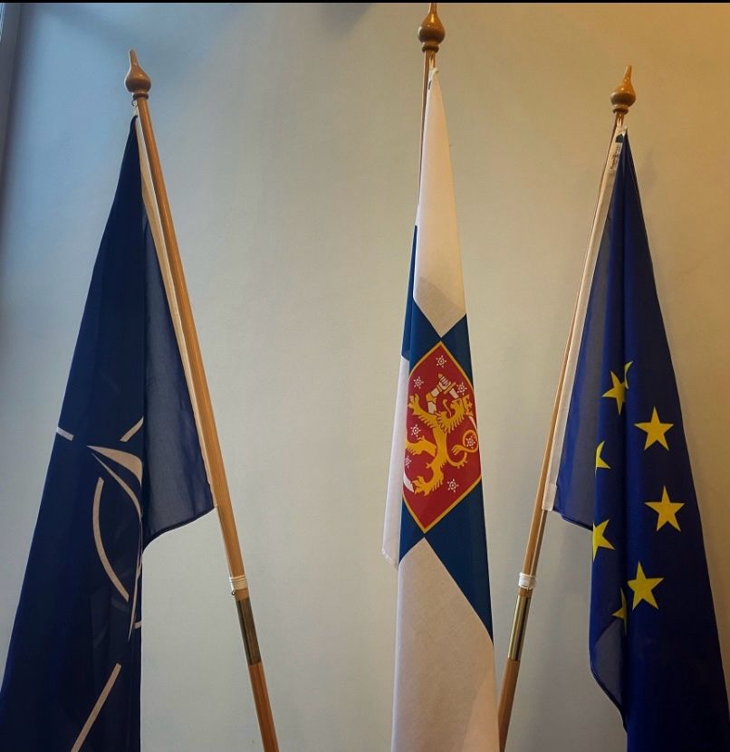 Kolme lippua salossa. Keskellä kielekkeinen Suomen valtiolippu (ent. Sotalippu), vasemmalla Nato-lippu ja oikealla EU-lippu.
