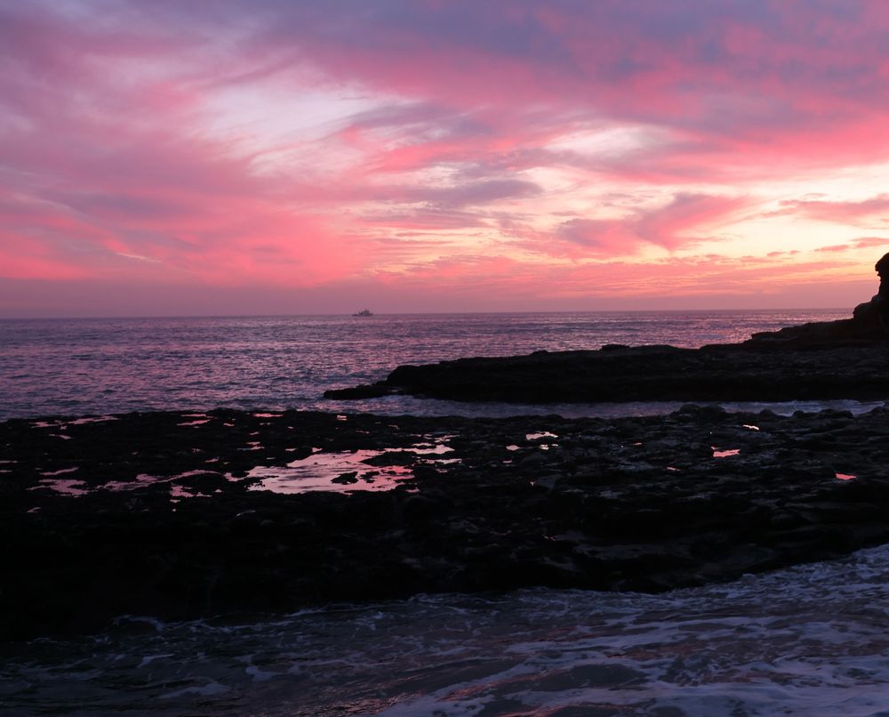 Bright pink cirrus clouds in sky reflected in tide pools on dark mudstone, Pacific Ocean on horizon.