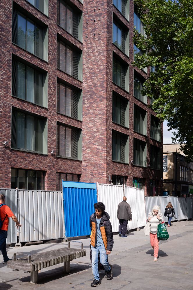 A street scene with a modern brick building in the background. The building has large windows and a facade made of red and brown bricks. In front of the building, there is a construction barrier made of white and blue panels. Several people are walking on the sidewalk, and there is a wooden bench in the foreground. The image captures an urban environment with ongoing construction or renovation work