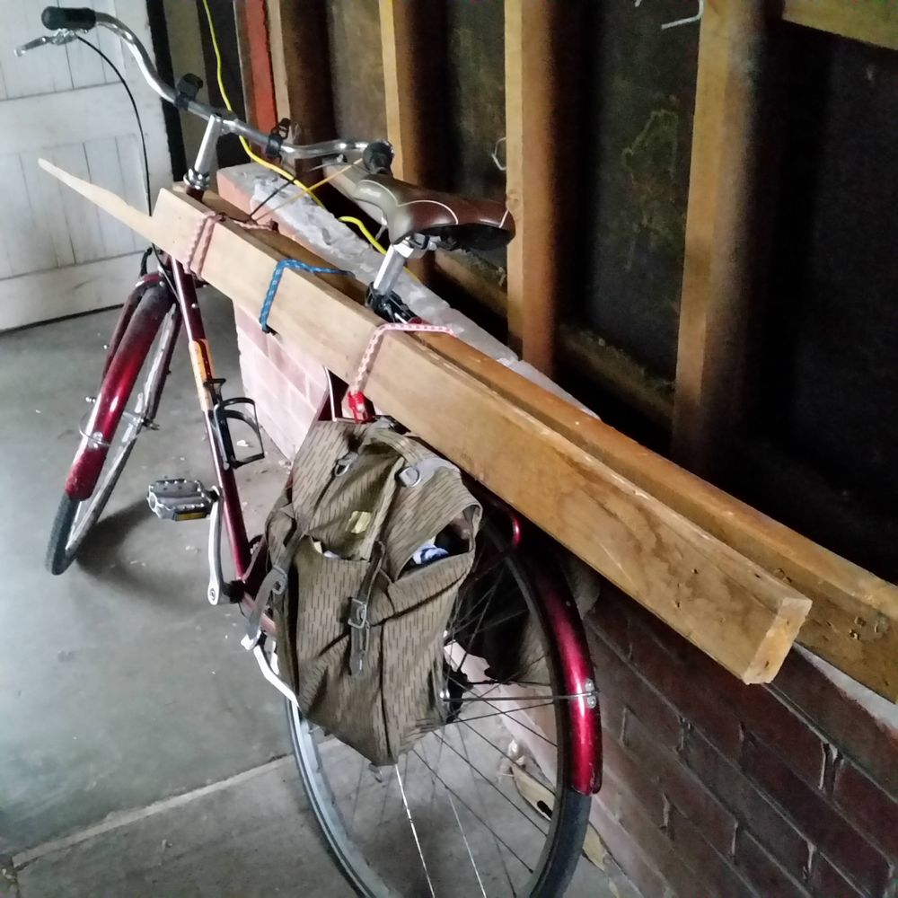 Red bicycle leaning against a wall, with heart rimu 4x2s strapped to it. 