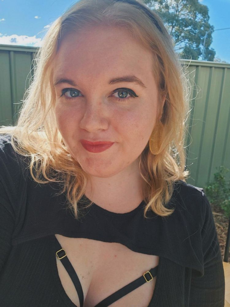 A selfie of a feminine looking person looking at the camera with a small close mouthed smile. They have wavy blonde, collarbone length blonde hair and green eyes. They are wearing a black shirt and large round gold earrings, as well as black eyeliner and red/pink lipstick.