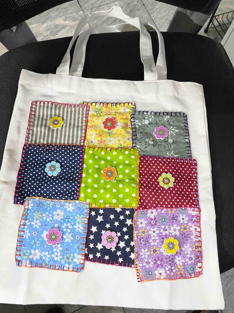 A shoulder bag with patchwork squared of different fabric, each square has a small flower in the middle.