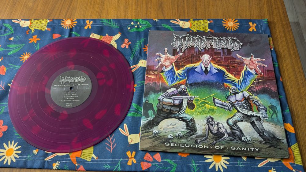 Picture of a vinyl record next to it's sleeve. The record is a translucent purple and the sleeve shows a puppet master pulling strings of two futuristic looking soldiers.

The album is Seclusion of Insanity by Mindfield, released in 2021. 