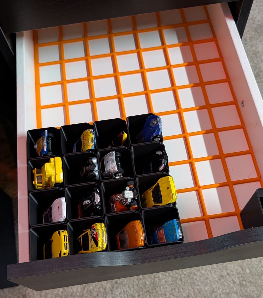 A picture of a pulled out IKEA Alex drawer with an orange grid and 15.5 3D printer cups, each with a car inside. 

The cars range from real to speed boat with wheels. There are more yellow cars than any other color. 