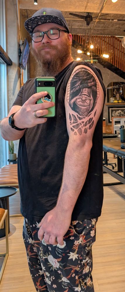 A man with an orange beard and glasses wearing a black T-shirt and flowery shorts. 
There's a tattoo of Frampt from Dark Souls 1 on his arm. 
Taken inside an industrial looking building. 