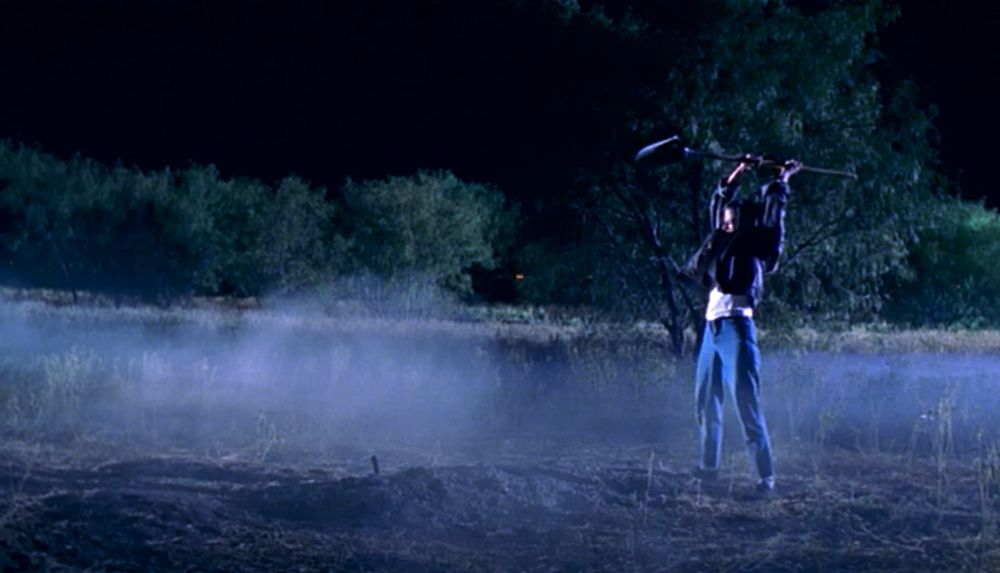 Debbie Rochon, now in a bomber jacket, holding a shovel aloft in both hands. She is standing of a fresh pile of earth and dust swirls around her 