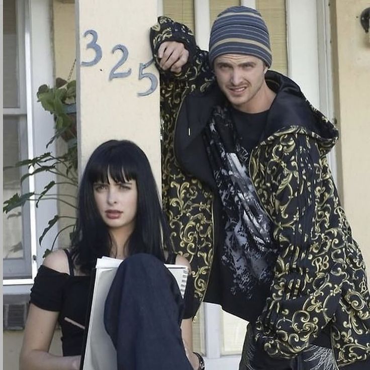 A still shot from the show, Breaking Bad, showing the characters Jesse Pinkman and Jane Margolis, with Jane sitting outside her apartment, with Jesse leaning on the pillar she is sitting against. They're both looking at something off camera.