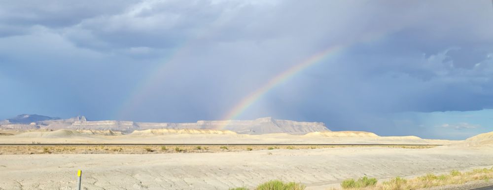 A double rainbow forms over a desert landscape, arching across an empty railroad track that stretches across the foreground. Stormclouds are in the distance over a wet mesa. No kings are in sight.