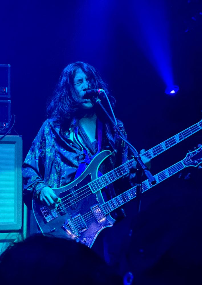 Takeshi from the band Boris, playing the bass of a double neck guitar. He is on a stage lit in blue