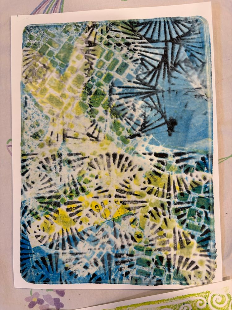 Gelli printed white rectangular paper with yellows, greens, and blues in the background. Black fan shapes in positive and negative forms are in the foreground.