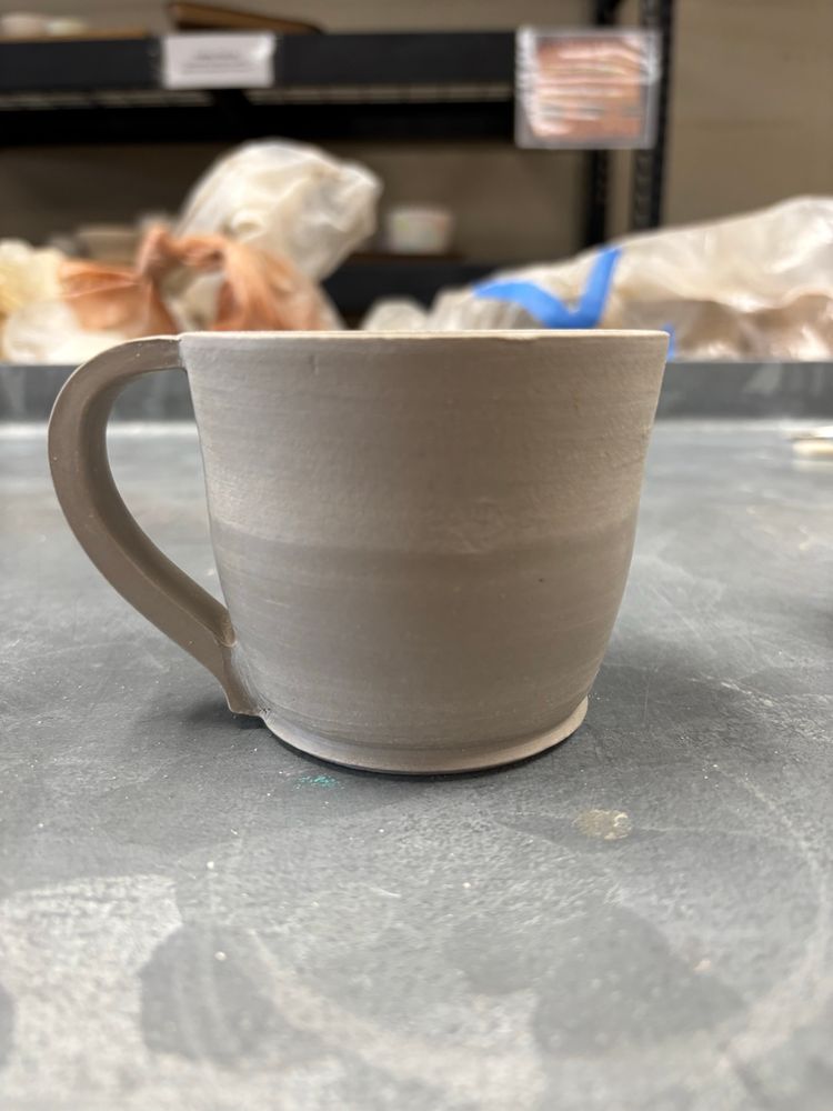 A small greenware coffee mug of light colored clay, with handle, sits on a grey table.