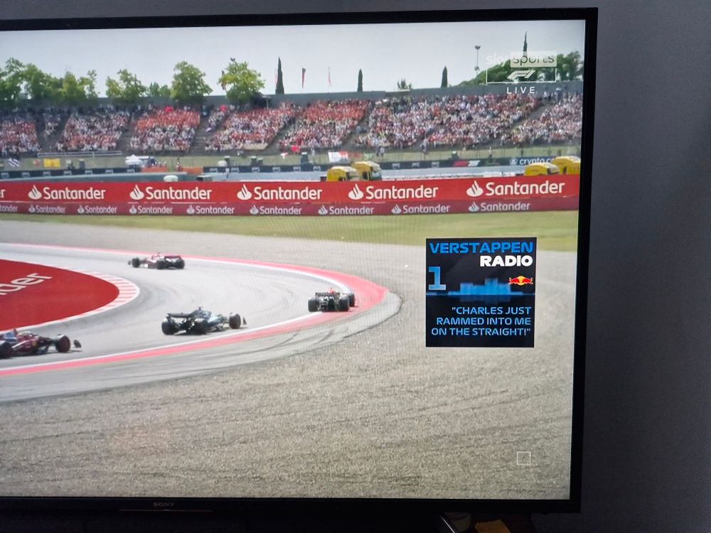 Formula 1 cars as photographed on a television screen. A team radio message from Max Verstappen reads: "Charles just rammed into me on the straight!" 