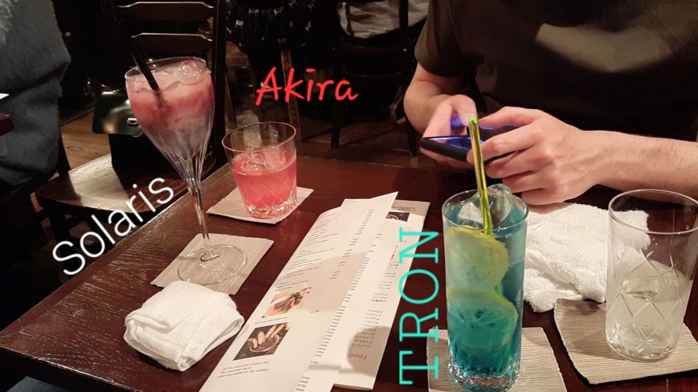 Three drinks on a table. An electric blue drink in a Tom Collins glass labelled TRON, a dark red drink in a wine glass labelled SOLARIS and a bright red cocktail in a tumbler called AKIRA. 