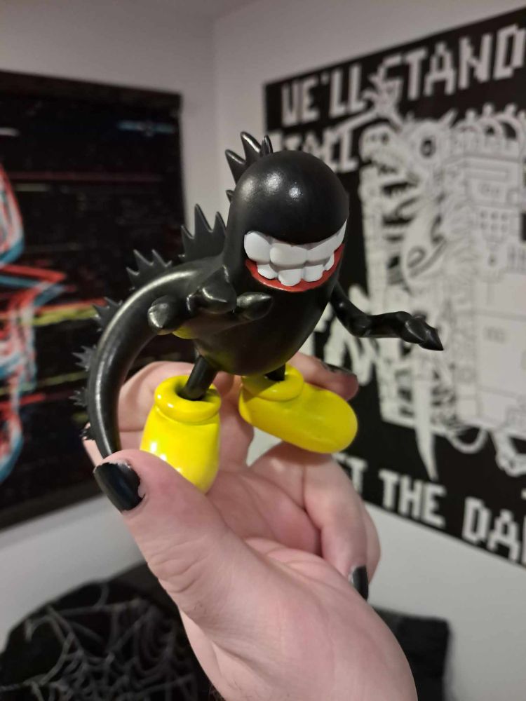 my hand holds up a 3.5 inch tall figurine. the body is a shiny black, constructed like Mickey Mouse's body (and wearing his signature round yellow shoes), but it has a long thick tail, with Godzilla's iconic back spikes running down it. the pill-shaped body doesn't have a distnct head - it just ends in an eyeless nub, with a big toothy grin that's just detailed enough that you can see the reds of the gums. a handpainted toy from kilrob's Legally Distinct collection.