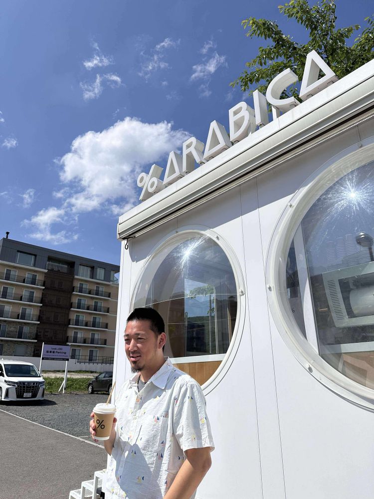 boso posing in front of a white coffee kiosk with circular windows. in the background is a parking lot and blue sky