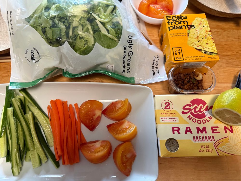 ingredients for 冷やし中華 laid out on the table. Ugly Greens salad mix, Sun Noodle Ramen Kaedama, cucumber, carrots, tomato, Just Egg folded, lemon, and some fried soy crumbles 
