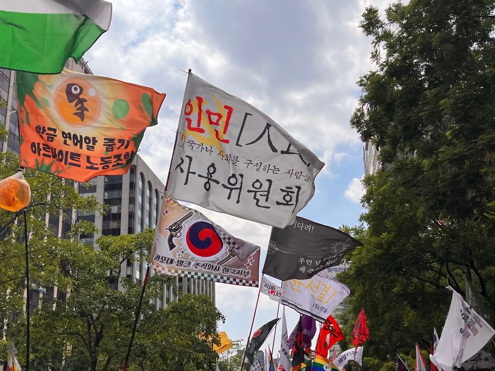 팔레스타인 연대 집회 현장의 사진. 맑은 하늘과 건물을 배경으로 여러 깃발이 휘날리고 있다. 깃발에 적힌 문구는 ’인민 사용위원회‘(국가나 사회를 구성하는 사람들), 카포네 밀크 주식회사 한국지부, 윤서결로 읽는 사람들, 황금 연어알 줍기 아르바이트 노동조합 등이 있다.