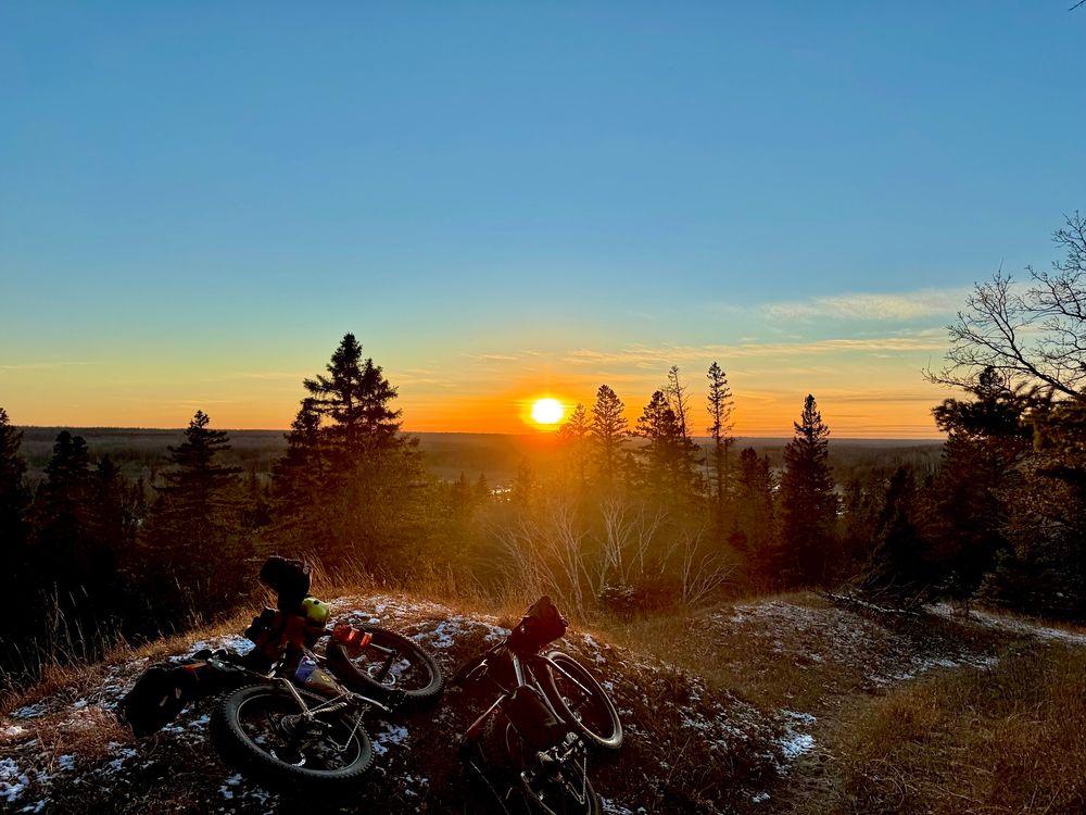 2 fat bikes laying on their side in the foreground with trees and and a sun in the background. Snow is dusting the ground.