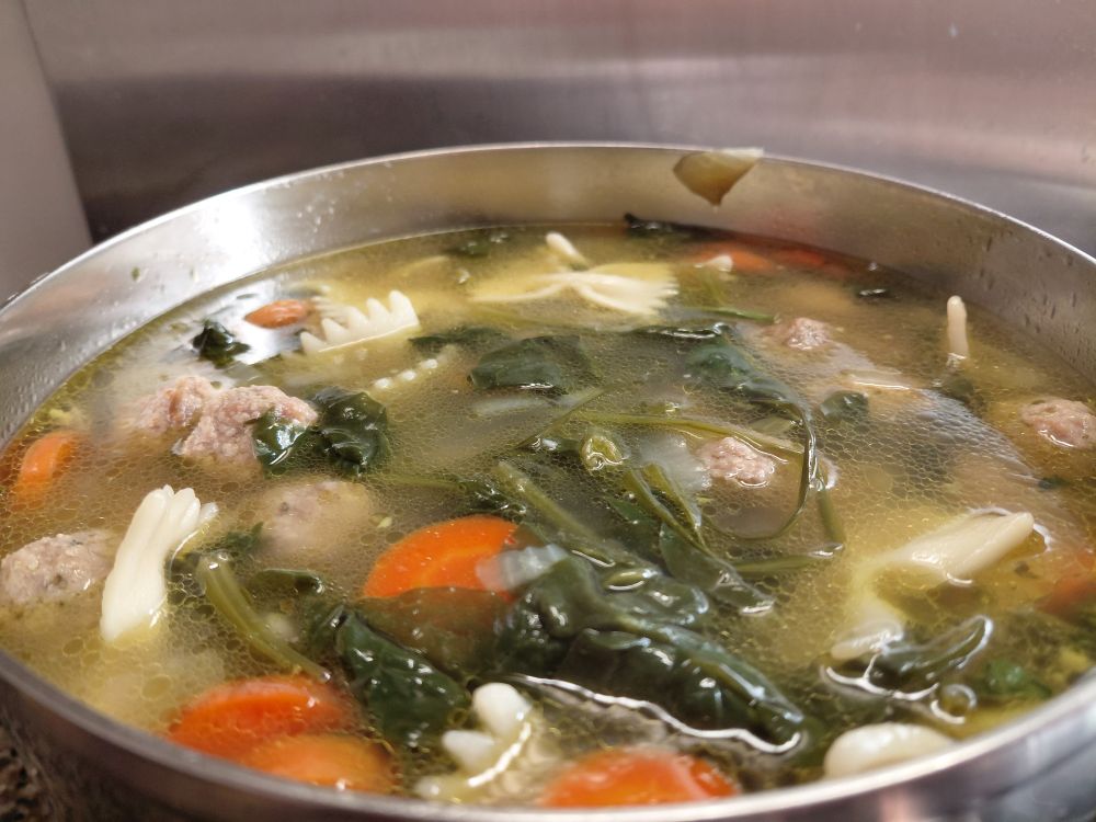 A close-up of a full soup pan showing broth, meatballs, carrots, spinach and pasta. 