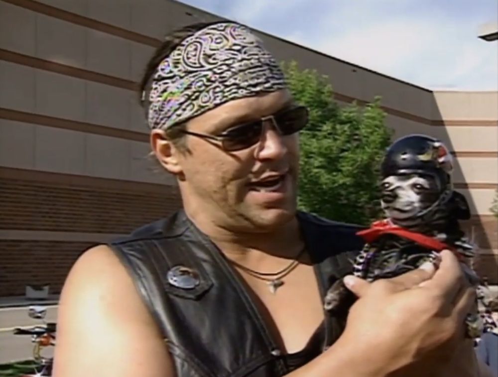 mongo standing outside getting ready to make the motorcycle ride to sturgis, mongo has a black leather biker vest on over a tank top and a black and white patterned bandana on, he's holding pepe who is wearing a harley davidson logo motorcycle helmet that is held on by what looks like a simple football grill, like a combo motorcycle football helmet, pepe also has a black biker jacket, red bandana, and metal chain to really complete the outfit