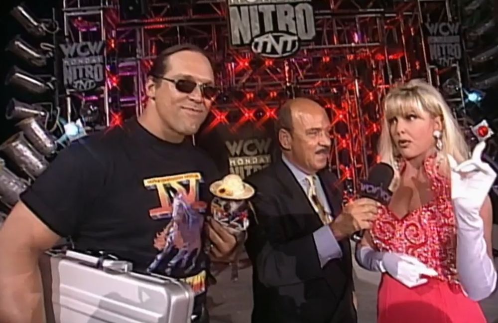 steve "mongo" mcmichael on the stage at wcw monday nitro wearing a 4 horsemen shirt and holding his haliburton briefcase under one arm, pepe is held by his other arm and is wearing a straw hat and colorful outfit like he's on vacation because these episodes were at disney world, to the right is mean gene okerlund with a microphone held up to debra mcmichael, who is wearing a gorgeous red evening gown, sparkly earrings, and long white gloves that go up to her elbow