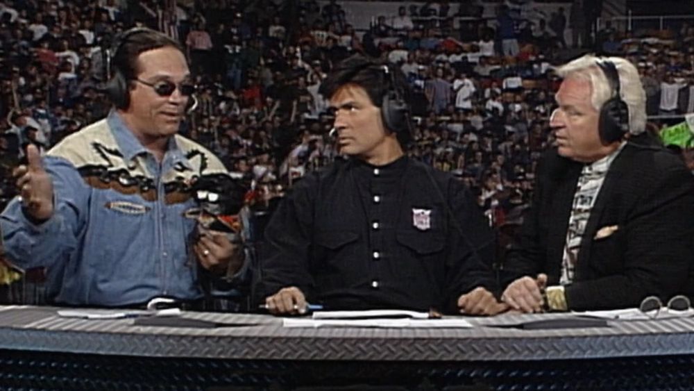the wcw monday nitro commentary desk, eric bischoff and bobby "the brain" heenan are looking over at mongo who is wearing a western style denim shirt with white/black/brown shoulder fabric highlights, mongo is holding pepe who has a black patterned outfit on, vibrant black sunglasses, and a truly absurd black haired wig that looks like someone emptied an entire can of hairspray on it