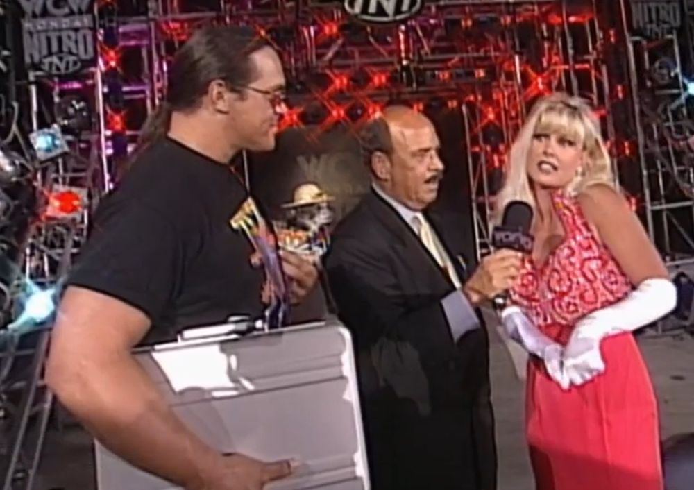 steve "mongo" mcmichael on the stage at wcw monday nitro wearing a 4 horsemen shirt and holding his haliburton briefcase under one arm, pepe is held by his other arm and is wearing a straw hat and colorful outfit like he's on vacation because these episodes were at disney world, to the right is mean gene okerlund with a microphone held up to debra mcmichael, who is wearing a gorgeous red evening gown, sparkly earrings, and long white gloves that go up to her elbow