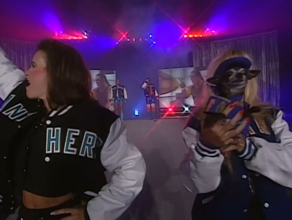 a close-up of debra mcmichael at the end of the ramp at wcw great american bash holding up pepe closer to the camera, pepe's outfit is difficult to see but he's wearing a blue chicago bears themed coat and hat, debra is in black shorts and a blue chicago bears letterman jacket with white sleeves, to her side is tara greene (wife of kevin greene) wearing black shorts and a black carolina panthers letterman jacket with dark gray sleeves, and tara is holding her fist up into the air, in the background behind them you can see kevin greene and steve "mongo" mcmichael emerging onto the stage about to head down the ramp