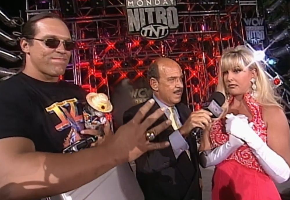steve "mongo" mcmichael on the stage at wcw monday nitro wearing a 4 horsemen shirt and holding his haliburton briefcase under one arm while also making a 4 horsemen hand gesture while showing off his super bowl ring to the camera, pepe is wearing a straw hat and colorful outfit like he's on vacation because these episodes were at disney world, to the right is mean gene okerlund with a microphone held up to debra mcmichael, who is wearing a gorgeous red evening gown, sparkly earrings, and long white gloves that go up to her elbow