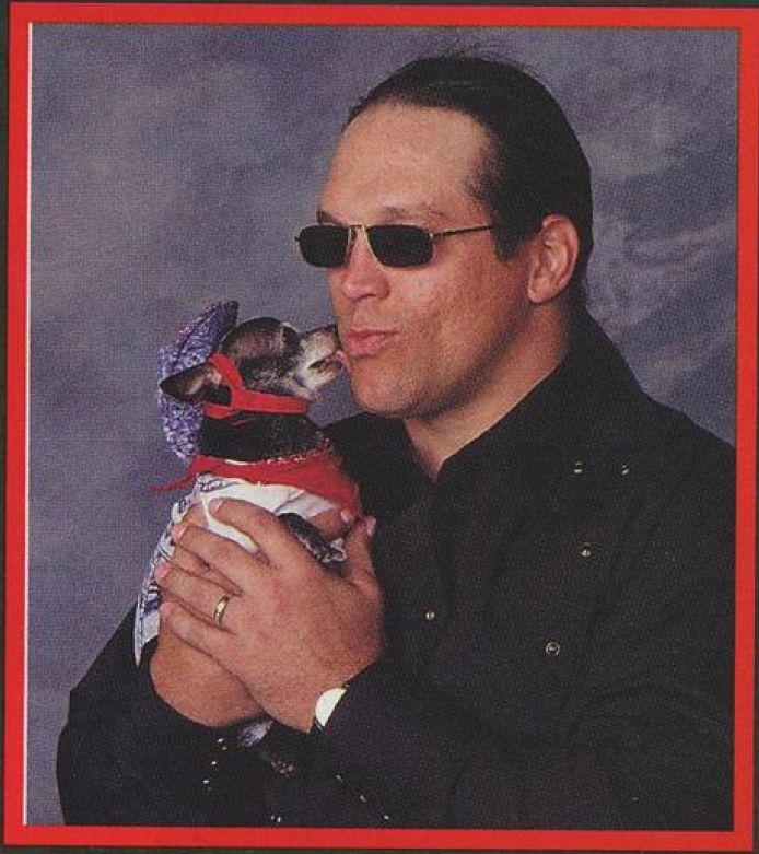 an image from the february 1997 issue of wcw magazine with steve "mongo" mcmichael in a black button-up holding pepe, pepe is wearing a white shirt, blue and white patterned cowboy hat, and his red bandana, pepe is turned to mongo and is licking his face