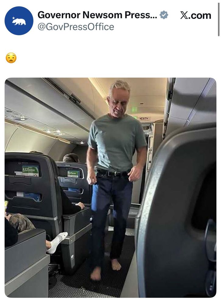 Photo of a barefoot RFK in the aisle of a plane wearing a t-shirt and jeans.