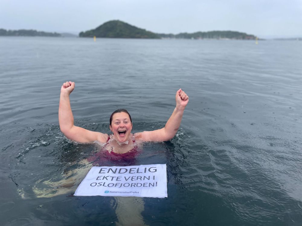 Pernille bader i Oslofjorden i regnet med en plakat foran seg: Endelig ekte vern i Oslofjorden! 