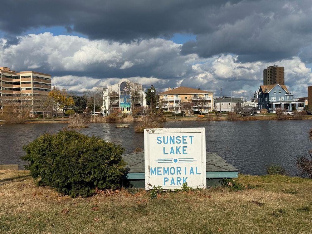 A sign that says "sunset lake, memorial park" in front of a lake with large beach houses and hotels in the background