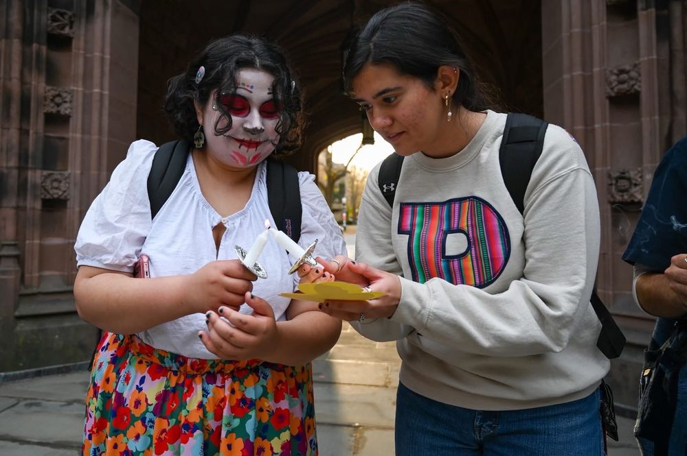 A student with sugar skull makeup shares her candle light with another student