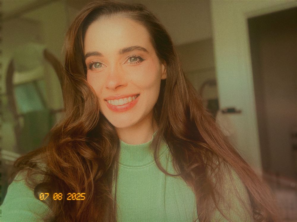 Seum, pale skin and long wavy brown hair and a long sleeve safe green mock neck on.