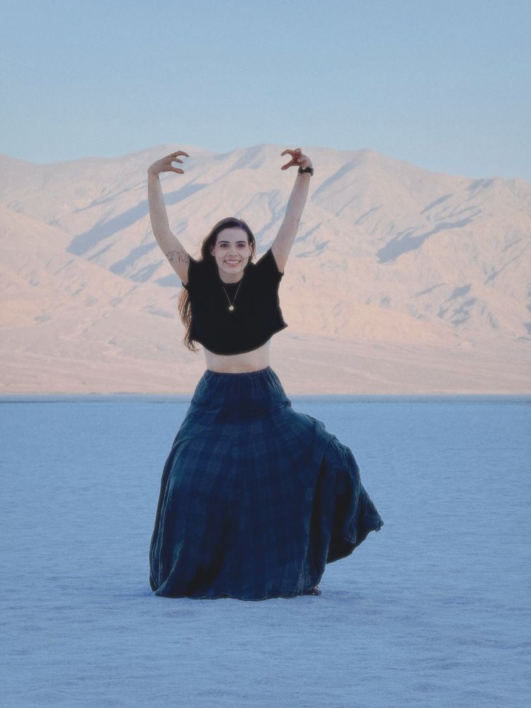 Seum, pale skin and long wavy brown hair, is wearing a black cropped tshirt, a gold circle pendant necklace, dark blue and green plaid maxi skirt with her arms in the air like a creature waiting to give you the worst high fives.

She is standing on the salted ground of Death Valley’s Badwater Basin at sunrise. 