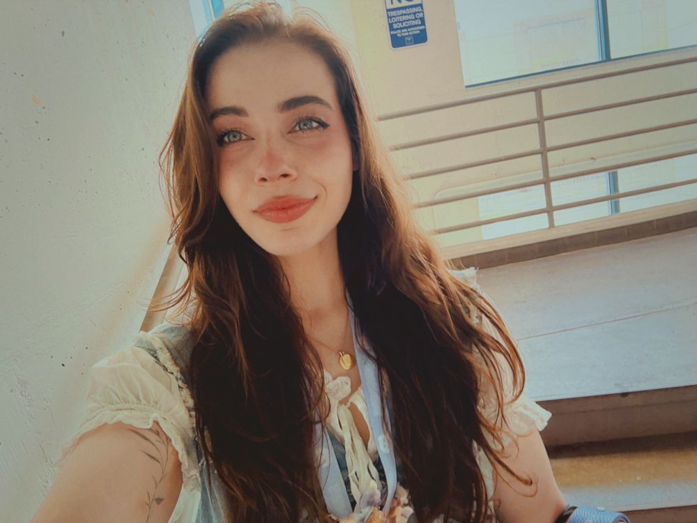 Seum, pale freckled skin with long wavy dark brown hair and light eyes, smiling wearing a delicate wait dress, blue lanyard, and taking a selfie in a stairwell.