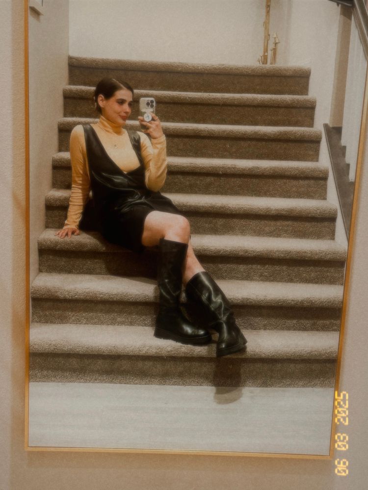 Seum, pale skin with brown hair pulled up into a hair clip, is wearing a yellow turtleneck shirt underneath a vegan leather dress hitting her mid-thigh. She has black leather tall boots on and is seated on her carpeted staircase.
