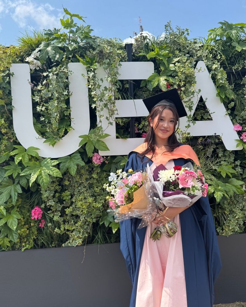 A graduate is standing in front of a UEA sign, holding flowers and smiling.