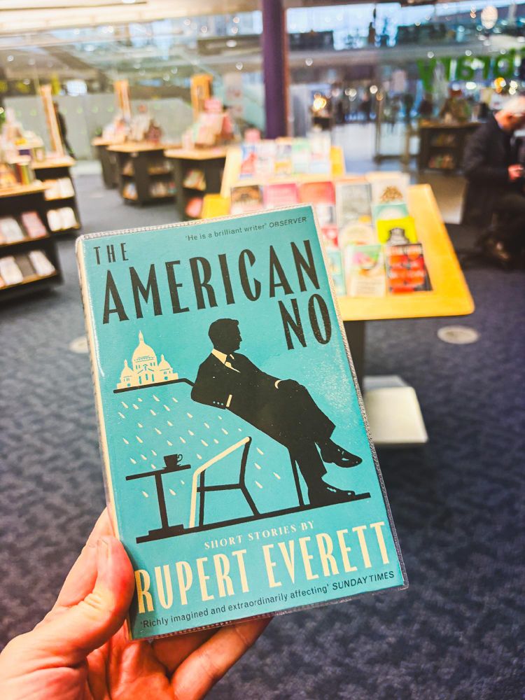 A hand holding a copy of "The American Novel: Short Stories by Rupert Everett" in a bookstore, with blurred shelves and patrons in the background.