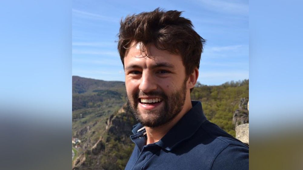 Person smiling in front of a scenic mountainous landscape.