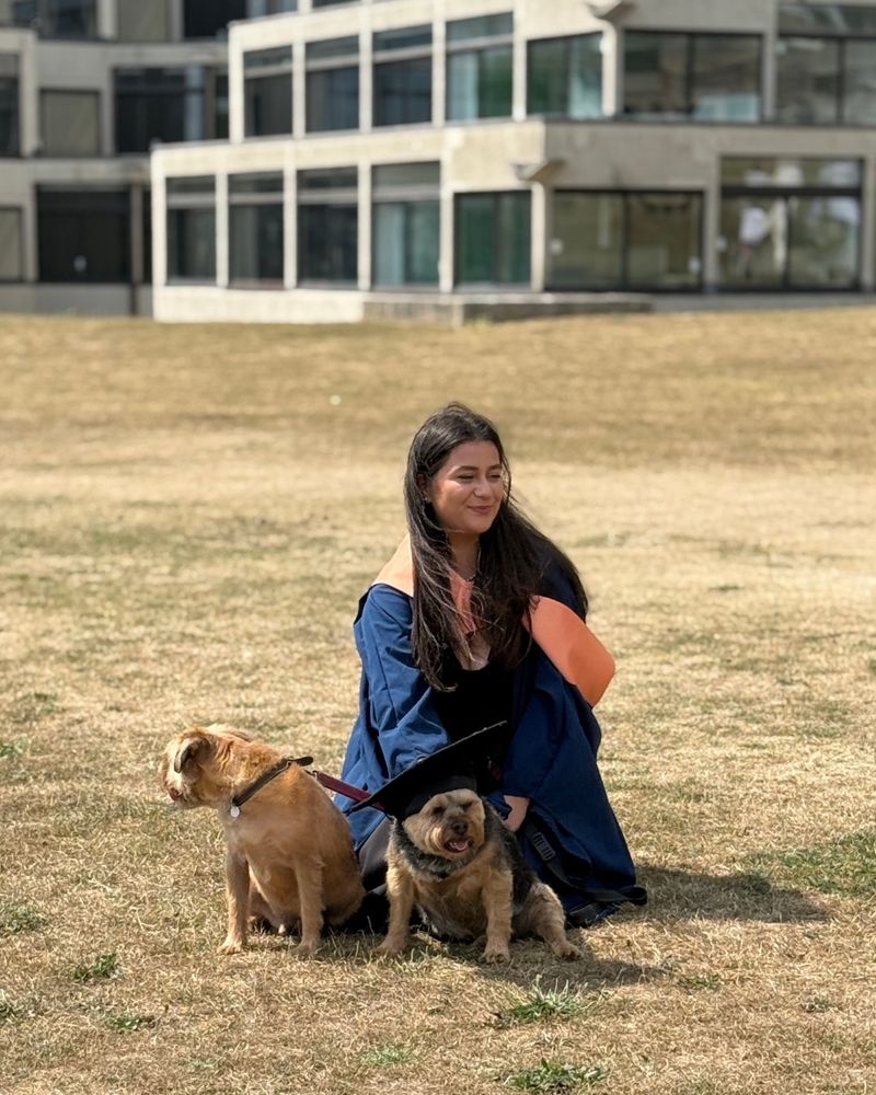 A graduate is kneeling down in front of the ziggurats with her two dogs. One of them is wearing her graduation cap.