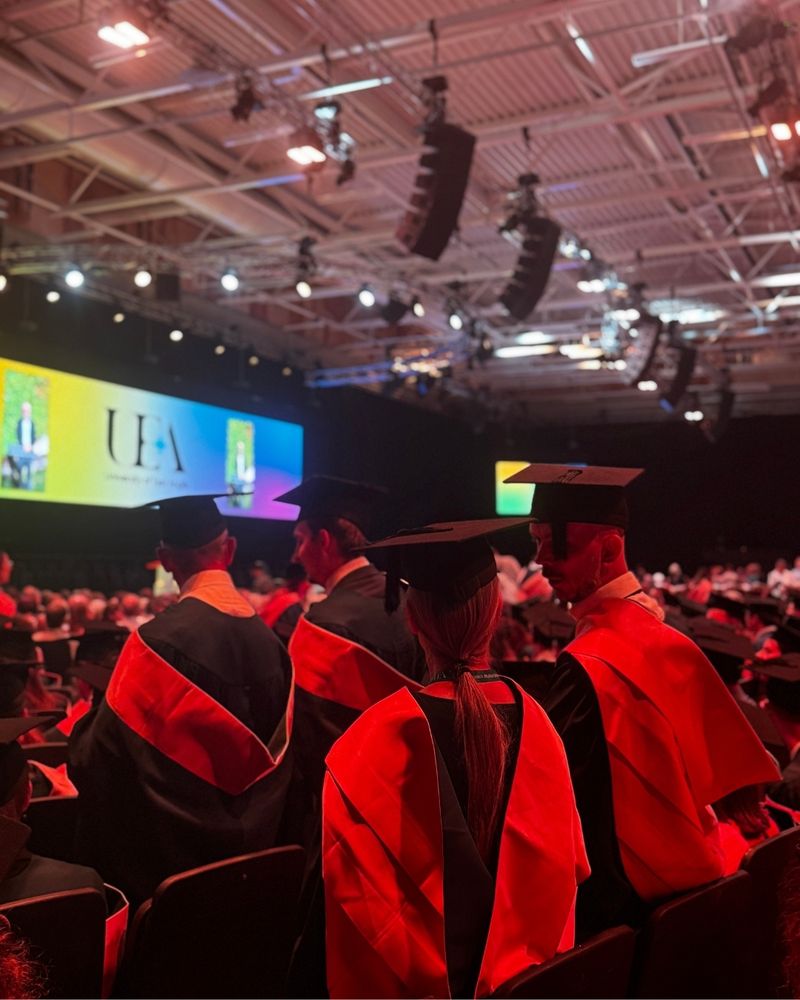 Four UEA graduates in caps and gowns are seen standing in the graduation hall, with the UEA sign in the distance.