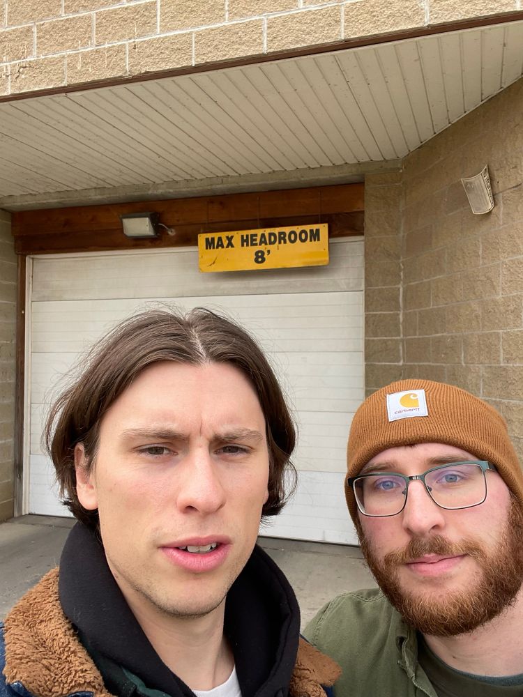 Two men stand in front of a docking bay door, with a sign that reads “max headroom 8’.”