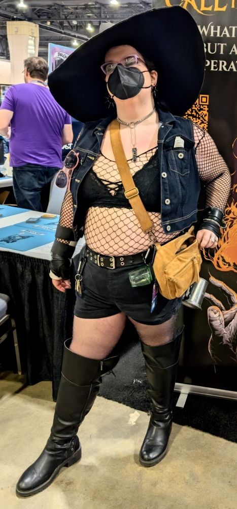 Florens stands in her knee high boots, star spangled stockings, short shorts, leather accessories, denim vest, comfy bra, fishnet top, witch hat and mask 